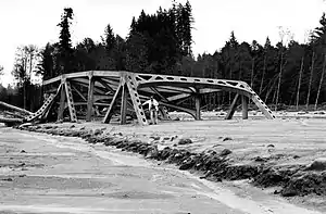 A broken steel bridge sits in a pile of mud, with a person posing next to a section that is buried halfway