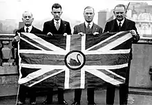 Black and white photograph of four men dressed in suits standing on a balcony holding a large flag depicting the Union Jack with a black swan in the centre.