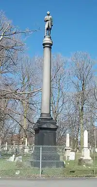 A tall stone monument behind a chainlink fence in a cemetery. A tall gray round column with a statue of a man atop sits on a darker pedestal