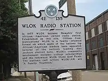 A white plaque with the designation 4E 135 at the top, reading "WLOK Radio Station: In 1977 WLOK became Memphis' first African-American owned radio station. Established on this site, WLOK is a family-oriented format on which many of the nation's top African-American leaders have appeared. Several of the nation's leading disc jockeys starred here. WLOK's community involvement includes college scholarships and the renowned WLOK Stone Soul Picnic. The station's call letters, WLOK-AM, are 'A Family Tradition.' "