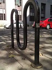 Ground-level view of a black-painted metal square tube bent into a curved M shape and bolted to concrete by a road. In the background are two cars parked in front of a long building.