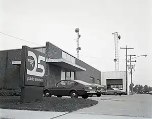 A one-story midcentury industrial building with a tower with antennae in the back. Visible are several 1970s cars as well as a sign with a stylized 25 and the words W V I Z TV, Public Television.