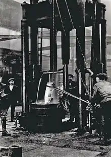 Workers stand around a large forge, where a shell burns red-hot.