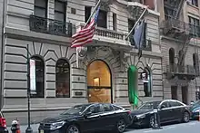 The ground story of the Mansfield Hotel as seen from across 44th Street. There are cars parked in front of the hotel. Behind the cars is a stone facade with an arched entrance and several windows.