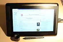 A monitor with the Dutch Wikipedia displayed. A large cable is plugged into the right side of the monitor. The Cintiq is standing upright on a desk, and lower down on the image is a digital pen and the pen holder.