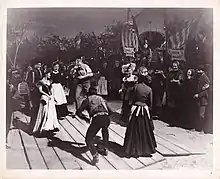 This is an outdoor scene with trees and the night sky visible in the background. In the foreground is a wooden dance floor consisting of long wooden boards butted roughly together. Four couples form the four sides of a square about 10 feet across. Two of the men are starting to move towards the center; all of the individuals are smiling and jovial. In the background there are about twenty more people watching, and behind them on the right there is a medicine show wagon. The wagon has several signs and banners. The topmost and largest reads "Dr. A. Locksley Hall"; smaller signs say "Hair Restored" and "Teeth Pulled". A man in a top hat is sitting regally on a small platform in front of the wagon. On the left are two musicians, one playing a flute and the other carrying a drum.