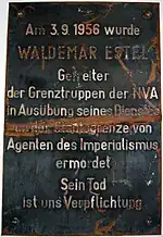 Damaged metal plaque reading "Am 3.9.1956 wurde WALDEMAR ESTEL Getreiter der Grenztruppen der NVA in Ausübung seine Dienstes an der Staatsgrenze von Agenten des Imperialismus ermordet. Sein Tod ist uns Verpflichtung."