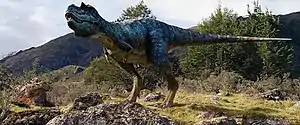 The Gorgosaurus, a bipedal dinosaur depicted with iridescent scales, stands on mostly rocky terrain with deciduous trees behind it.