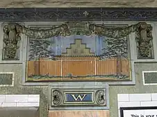 A faience mosaic on the wall that depicts a New Amsterdam step-gabled house with the palisade wall in front of it. The mosaic is topped by blue faience swags. There is a faience cornice with scrolled and foliate detail.