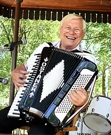 Seated, smiling man with wire-frame glasses, playing a piano accordion that is decorated with the name "OSTANEK".