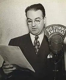 Black and white photograph of a male wearing glasses and standing next to a microphone