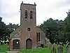 A small brick-built church with the tower in the centre. To the right are gravestones and in the background are trees