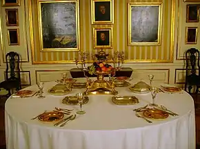 A round table covered with white table cloth and set for six with silverware and a centrepiece, standing in front of a wall covered with yellow and white striped wallpaper and framed paintings