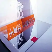 An orange sign with the band's name, being reflected through two different mirrors, one on the floor and the other leaning against a wall