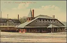 Postcard showing a two-story railroad station with a smaller second story. The roof from the second story continues down to the ends of the first story. A small turret is located on one end.