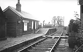 A small wooden hut labelled "Westcott". In front of the hut is a deserted low wooden railway platform with a short section at a much greater height; the only objects on the platform are three large lamps. A single railway track leads past the platform; the line branches immediately past the end of the platform. A cat is asleep on the railway track.