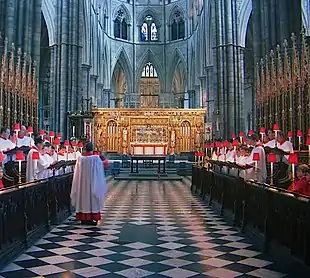 The choir master walking down the aisle of the abbey, with choirboys in red and white robes standing in stalls down its length.