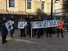 Demonstrators at a solidarity rally in Scotland with a banner painted with the words "We stand with the Wetʼsuwetʼen".