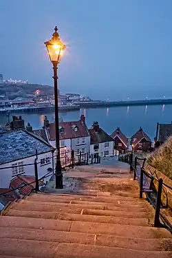 A twilight image of a steep staircase descending, with an old-style gas lamp lit beside it, with a harbour, headland and sea beyond