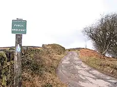 A moorland road with a bridleway sign next to it, and a long sandstone wall on the left side