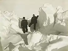  Two men in heavy clothing stand surrounded by mounds of ice which extend well above the height of their heads