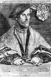 A middle aged man, Wilhelm, Duke of Jülich-Cleves-Berg, is sitting at a table. He is dressed in a soft cap that falls to the side of his head. He is wearing fur-trimmed robes, and nestled in the edges of the robes is a chain, with a cross at its nadir. His hands are folded on the table before him. He is wearing several rings, and one hand holds a pair of gloves. The family crest hangs on the chair behind him.