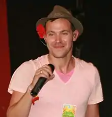 A man with a brown cap wearing a pink T-shirt and holding a microphone. He is looking to the left and smiling.