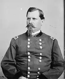 Black and white photo shows a stern-looking man with a moustach and pointy beard. He wears a dark military uniform with two rows of buttons.