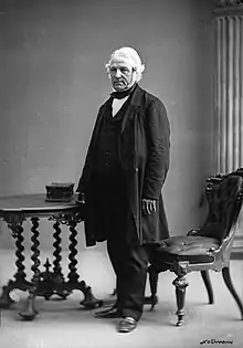 A man aged around 60 with thick white hair and a heavy black suit stands between an ornate wooden chair and a table.