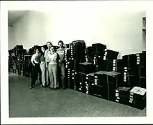 A group photo of 5 herbarium employees, standing in front of a large pile of specimens in boxes