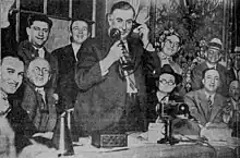 Black and white photo. William Hale Thompson stands before a desk, surrounded by others, while using a telephone of the candlestick telephone-design, with the mouthpiece held to the right-side of his face by his right hand and the earpiece held to his left ear by his left hand