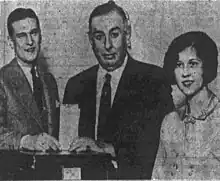  Black and white photograph. Thompson stands before a ballot box, inserting his ballot paper into the box. He has a male. To the left of Thompson is a male, and to the right of him is a woman, both also standing