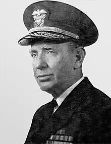 Head and shoulders of man in dark suit and tie wearing white peaked cap with gold braid. He has several rows of ribbons on the left breast.