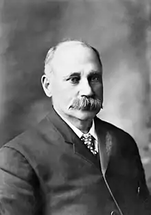 A white man with receding light hair and a thick grey moustache, wearing a suit