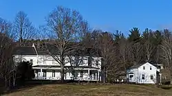 A long two-story white house with a black side-gabled roof and a tall bare tree in front of it seen from a distance. Next to it a much smaller side-gabled house, similar in architectural style, is seen from the side.