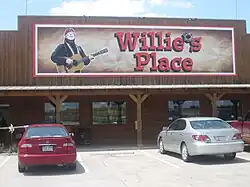 A store with a sign that reads "Willie's Place". The apostrophe is replaced in the sign by a bullet hole. The structure of the store is constructed in wooden with three columns. There are four windows and there are a red and a grey car in the parking lot.