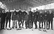 Black and white photo of eight hockey players in equipment and uniforms standing on an ice rink, with four men in suits and ties
