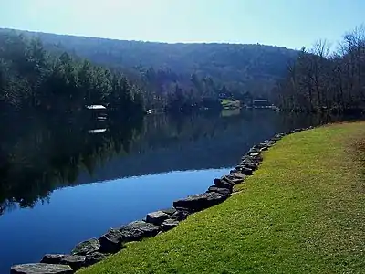 A small lake with a smooth surface reflecting the blue sky above and a hill beyond it with some buildings along the far shore. In the foreground, at lower right, the edge is marked by a stone retaining wall