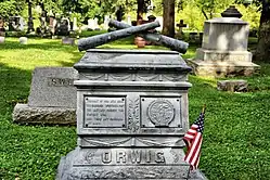 Grave marker with crossed cannons on top, other graves in background.