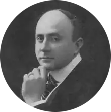 head and shoulders of balding white man in early middle age, clean-shaven, in lounge suit and tie, semi-profile, looking towards the camera
