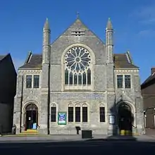 Front view of a pale stone building dominated by a large rose window sitting on six narrow windows of varying height. The main body, with the rose window and two spirelets, is flanked by identical recessed sections with round-arched entrances and paired rectangular windows.