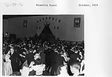 A dance hall full of young students, with a tipi on one wall below the word Wuaneita