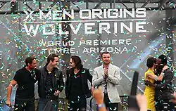 Atop a stage are three man in black clothing, Liev Schreiber wearing a gray jacket and black pants, and Lynn Collins, wearing a yellow dress, hugging will.i.am, who is in black clothing. In the background is a billboard reading "X-Men Origins Wolverine: World Premiere – Tempe, Arizona. Colored paper flies through the stage.