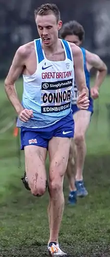Man in white top and blue shorts running, with mud on his legs.