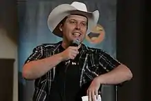 Man in cowboy hat holding microphone and leaning on microphone stand.