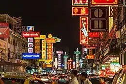 A street during sunset lined with many stalls and shops with a lot of signs bearing Thai and Chinese names