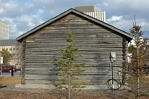 Back of the building, with no windows but an electric meter on the right and a small evergreen tree in the center
