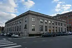 A tall light-colored square two-story stone building with a balustrade and pilasters on the front on an urban street. An American flag flies from a pole on the right. Across the top of the building the words "United States Post Office" is engraved.