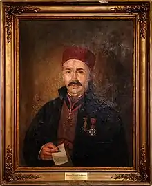 Portrait of a decorated and mustached older man wearing a fez. He faces the onlooker and holds a piece of parchement.