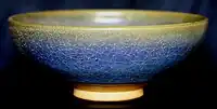 A ceramic bowl is viewed from one side. The glaze has a crazed and mottled finish, varying from yellow at the rim of the bowl to blue below.